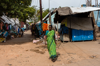 Une femme en sari vert longe une rangée d'habitations modestes couvertes de tuiles et de bâches.