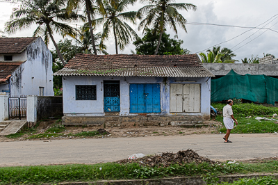 Un homme longe une maison aux volets bleus sous les cocotiers.