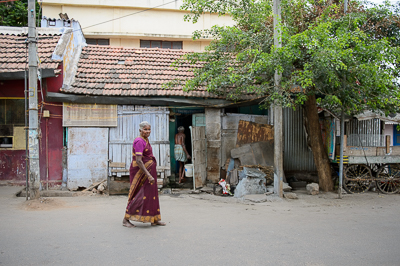 Une femme âgée en sari violet traverse la rue devant des maisons aux toits de tuiles.
