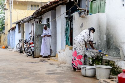 Une femme lave du linge au point d'eau installé devant les maisons, sous le regard d'un homme âgé.