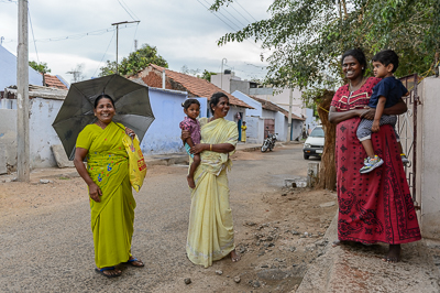 Trois femmes, dont deux portent un enfant, sourient sous un grand parapluie noir.