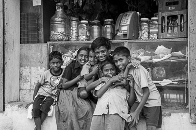 Six enfants se serrent en riant devant la vitrine d'une petite échoppe, dans un portrait en noir et blanc.