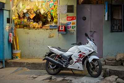 Une moto blanche stationne devant une petite épicerie vivement éclairée et couverte de marchandises.