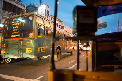 Dans la circulation nocturne, un bus est cadré entre le rétroviseur et la structure du rickshaw.