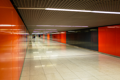 Les parois alternent le rouge vif et le noir brillant, formant une sorte de code visuel très net. Le plafond quadrillé et les néons alignés accentuent encore la perspective. Le sol poli renvoie des reflets qui dédoublent l’espace. On a l’impression d’entrer dans un décor futuriste, presque abstrait, où les rares silhouettes humaines deviennent des repères minuscules.
