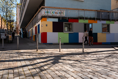 Leonhardskrippe, Stuttgart.
Ce qui attire d’abord l’œil, ce sont ces pans de murs colorés qui rappellent des cubes de jeu ou un dessin d’enfant. Le contraste est fort avec la rigueur du bâtiment au-dessus, gris et anguleux. Le pavé au premier plan, les ombres des arbres et les passants donnent un ancrage quotidien, un côté « petite scène de quartier ».
