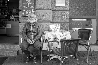 Assis bien droit devant une table encore vide, il a des allures de patriarche urbain. Sa veste matelassée et sa barbe blanche contrastent avec la nappe bariolée “Coffee” et l’ardoise qui vante mortadelle et végétarien, sous le regard goguenard de Mario. Stuttgart lui offre un trône improvisé, simple mais digne.