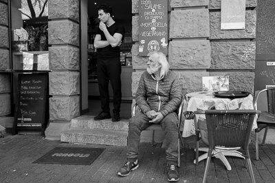 Le jeune serveur, bras croisés, observe la rue, concentré, presque soucieux. À ses pieds, l’homme plus âgé s’est assis, immobile, le regard porté ailleurs. Entre eux, aucune parole : juste la façade rugueuse, la nappe imprimée et le panneau vantant panini et mortadella. Deux vies en suspens, réunies par hasard sur le même seuil de Stuttgart.