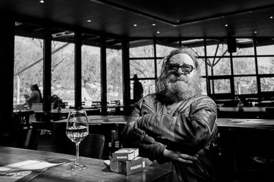 Dans la lumière dure de l’après-midi, il trône comme un personnage de roman. Lunettes noires, barbe en cascade, veste de cuir usée, bras croisés : rien ne peut l’atteindre. Sur la table, un verre de vin blanc et deux paquets de Gauloises plantent le décor d’une résistance tranquille. Derrière lui, les silhouettes passent, indifférentes. Lui, il pose sans poser, icône intemporelle d’un Stuttgart de café et de fumeurs.