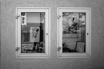 Dans la première fenêtre, un visage de magazine promet la coupe parfaite, stylée, moderne. Dans la seconde, le reflet du trottoir, un menu encadré, un bric-à-brac végétal. Chez ce coiffeur de Stuttgart, la vitrine hésite : rêve de beauté à gauche, réalité du quotidien à droite.