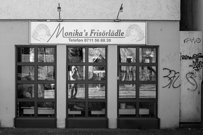 Devant le salon de coiffure de Monika, les reflets font tout le travail : les silhouettes de clients, la rue, les graffitis, et soudain l’apparition du photographe. L’appareil porté à l’œil, il s’invite dans la scène sans y entrer vraiment. À Stuttgart, le coiffeur devient studio improvisé et la vitrine miroir d’un autoportrait furtif.