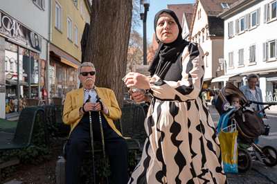L’homme à la veste jaune trône, immobile, ses bâtons plantés comme un sceptre. La femme passe devant lui, robe ondulante, gestes rapides. Dans le fond, l’enfant observe. Trois présences, trois rythmes. La rue compose sa propre mise en scène.
