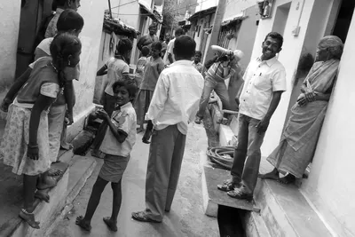 En noir et blanc, je photographie dans une ruelle où des enfants et des adultes se tiennent devant les maisons, de part et d’autre d’un caniveau.