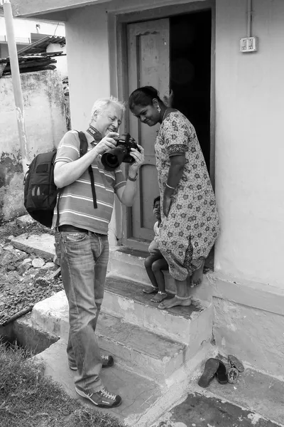 En noir et blanc, je montre la photographie sur l’écran de mon appareil à une jeune femme sur le seuil d’une maison ; un enfant est assis derrière elle et des sandales reposent sur la marche.