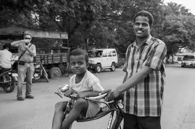 En noir et blanc, un homme souriant tient le guidon d’un vélo sur lequel un enfant est assis ; un autre photographe apparaît à gauche, en arrière-plan.