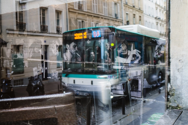 Reflet d’un bus aux contours turquoise confondu avec une vitrine de portraits, dont plusieurs visages semblent assis derrière les fenêtres