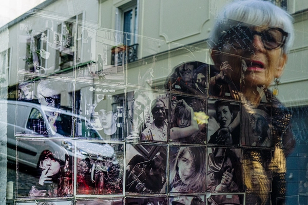 Vitrine couverte de portraits photographiques sur laquelle se superpose le reflet en gros plan d’une femme aux cheveux blancs et aux lunettes noires