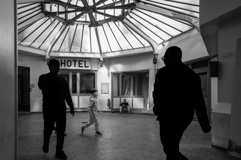 Sous la verrière du passage du Prado à Paris, un enfant traverse entre deux silhouettes sombres ; au fond, un homme est assis près de l’entrée d’un hôtel.