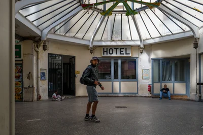 Un homme coiffé d’un chapeau, portant un sac à dos et un short, se tient sous la verrière devant l’hôtel du passage du Prado.