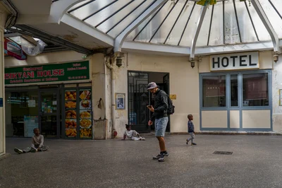 Sous la verrière, un homme au sac à dos regarde vers ses mains tandis que des enfants occupent l’espace devant l’hôtel et un restaurant.