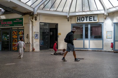 Un homme au sac à dos marche devant l’hôtel ; des enfants se déplacent autour de lui sous la verrière.