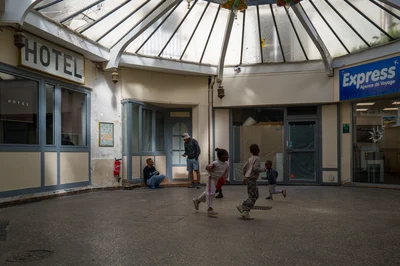 Des enfants traversent l’espace entre la façade de l’hôtel et la boutique à l’enseigne Express ; un homme est assis au pied d’une fenêtre.