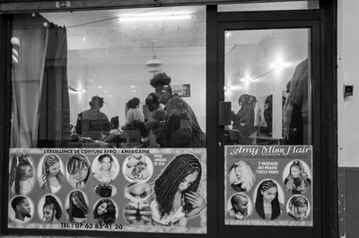Photographie en noir et blanc d’un salon de coiffure vu depuis le passage, avec des personnes à l’intérieur et des portraits de coiffures sur la vitrine.