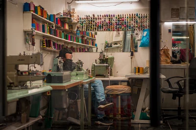 Un atelier de couture vu depuis le passage, avec une machine à coudre, des rangées de bobines colorées et une personne assise au fond.