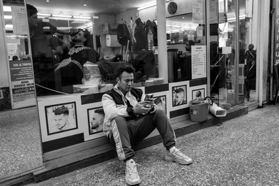 Photographie en noir et blanc d’un homme assis devant la vitrine d’un salon de coiffure, regardant un téléphone tenu dans ses mains.